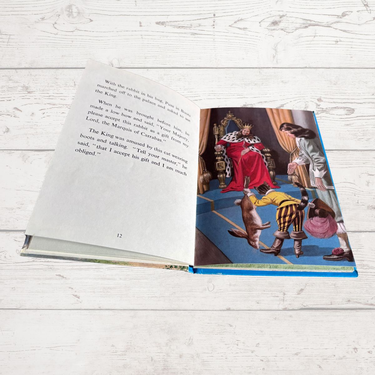 An open children's book lying on a white wood surface, showing printed text on the left page and a colorful illustrated scene on the right page with a king in red robes and a woman with a child.