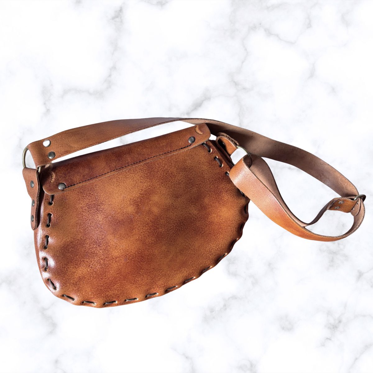 A brown leather shoulder bag with a curved, saddle-like shape, hand-stitched edges, and a matching adjustable leather strap, displayed on a white marble background.