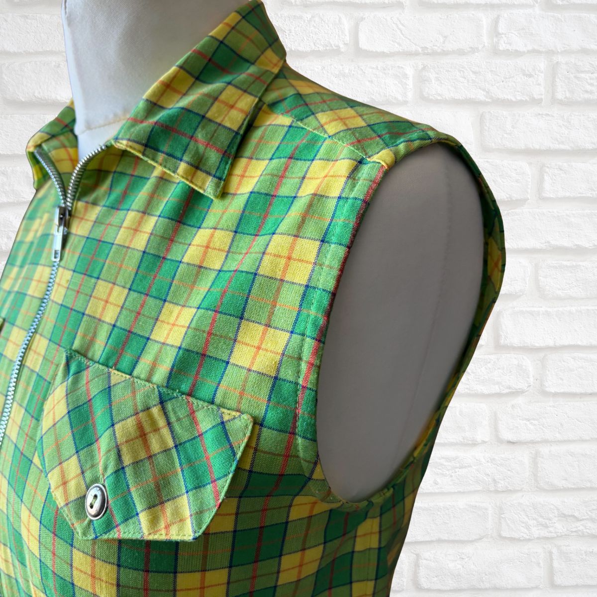 Green and yellow plaid vest on a mannequin against a white brick wall.