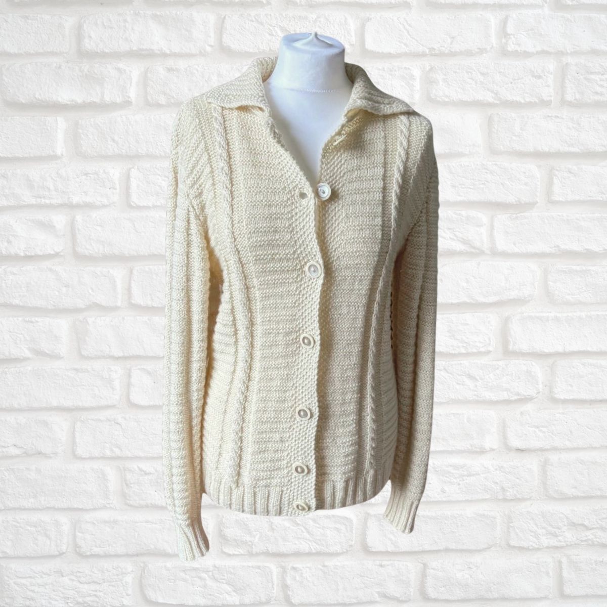 Cream cable-knit cardigan sweater with a collar and button-front closure, displayed on a white mannequin torso against a white painted brick wall.