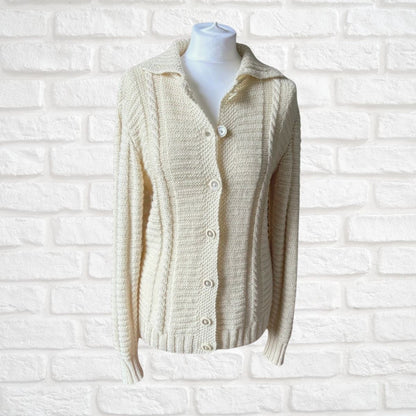 Cream cable-knit cardigan sweater with a collar and button-front closure, displayed on a white mannequin torso against a white painted brick wall.