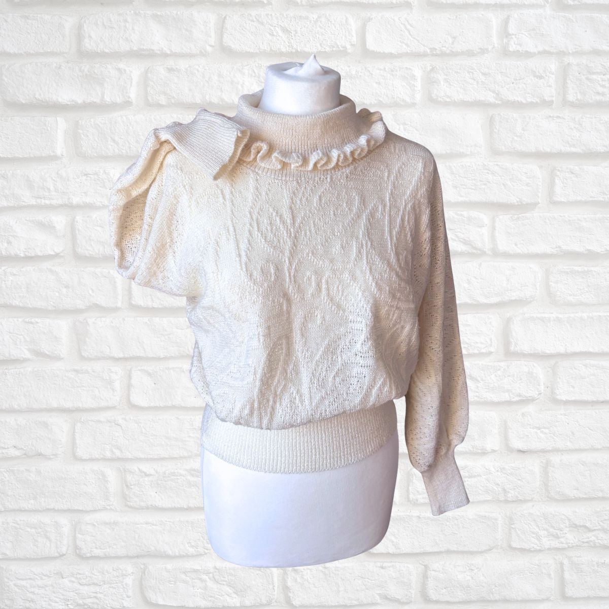 Cream-colored textured knit sweater with a ruffled mock neck and puff sleeve, displayed on a white mannequin torso against a white brick wall background.