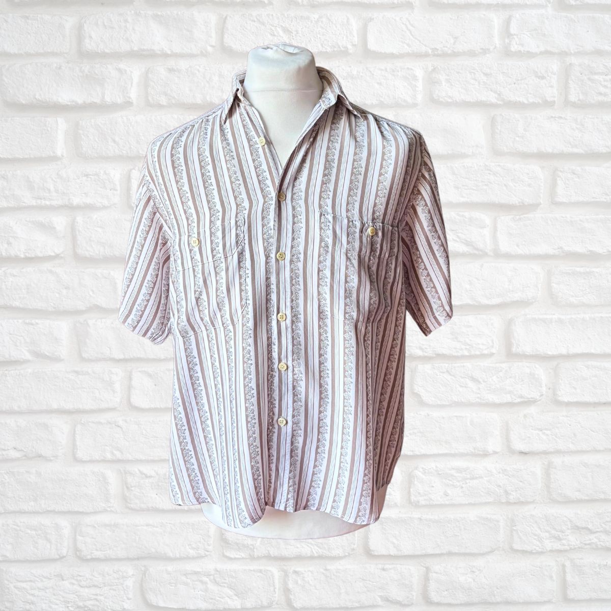 A short-sleeve button-up shirt with vertical textured stripes in beige, brown, and white, displayed on a white mannequin torso against a white brick wall background.