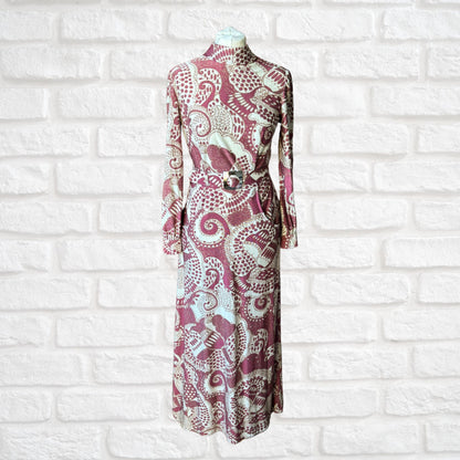 A vintage 70s long-sleeved maxi dress in a cream, burgundy, and brown abstract print, with a high turtle neck, long sleeves with silver button fastened cuffs, and a matching belt, displayed on a mannequin against a white brick wall background.