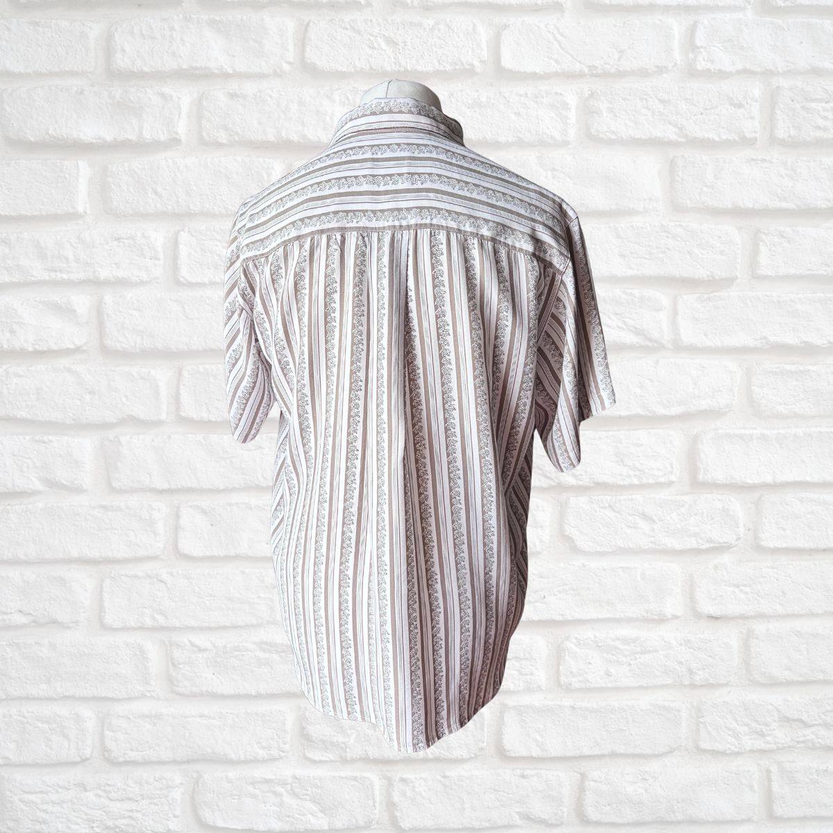 A short-sleeve button-up shirt with vertical textured stripes in white and gray, displayed on a mannequin torso against a white brick wall background.