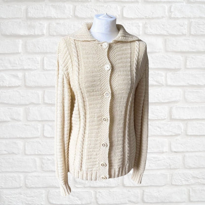 A cream-colored cable-knit cardigan sweater with a collar and button-front closure, displayed on a white mannequin torso against a white painted brick wall.