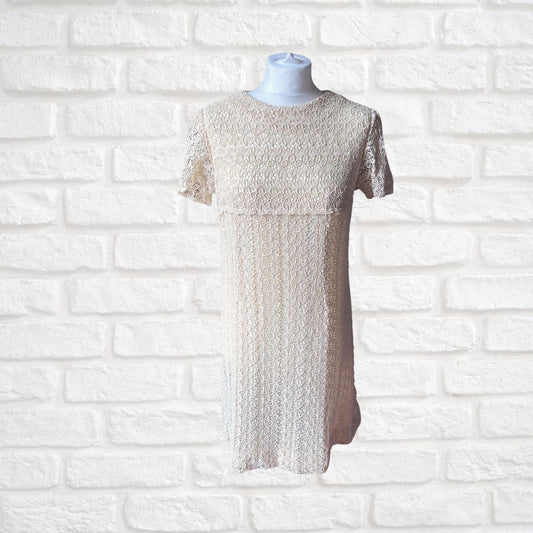 A short-sleeve lace dress in a light beige or cream shade, displayed on a white mannequin torso against a white painted brick wall background.
