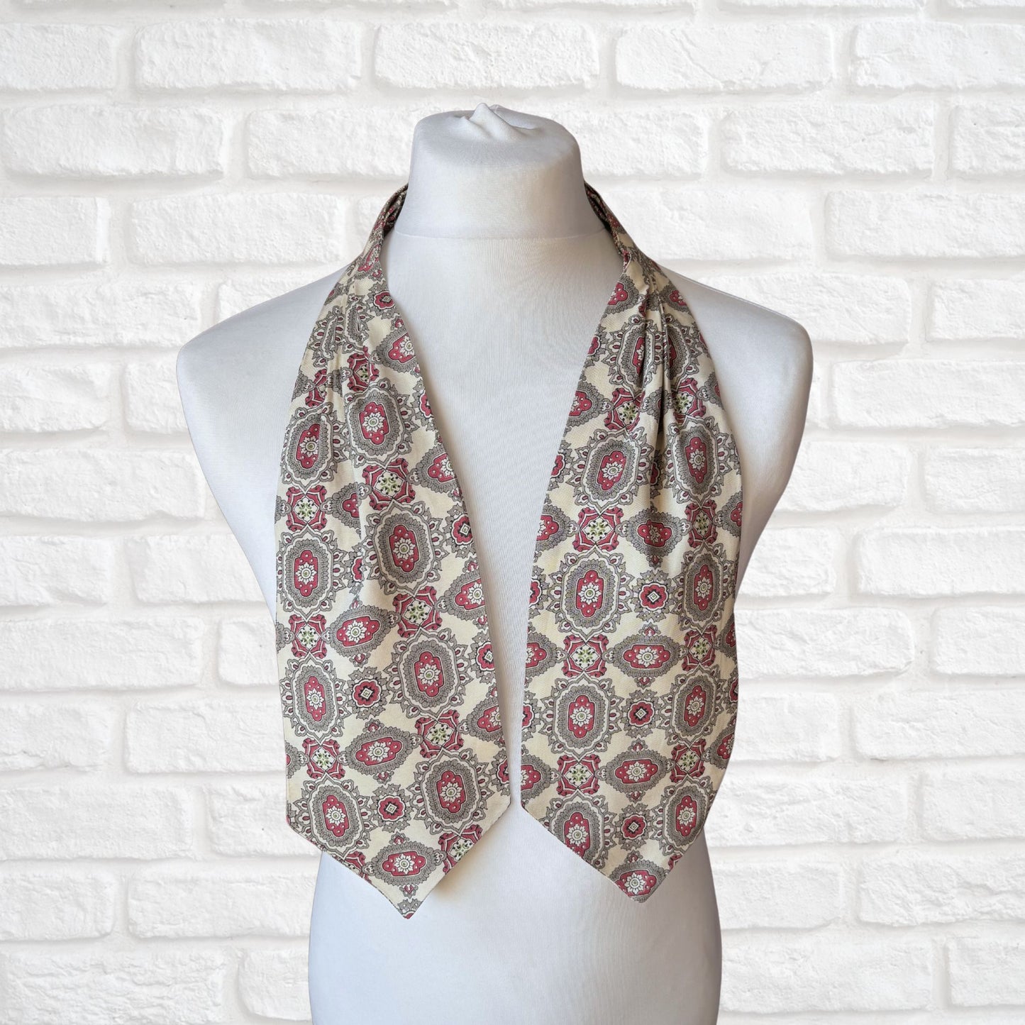 A vintage 60s mod style cream cravat with a paisley print featuring baroque style red, grey, and green patterns, displayed on a mannequin against a white brick wall background.
