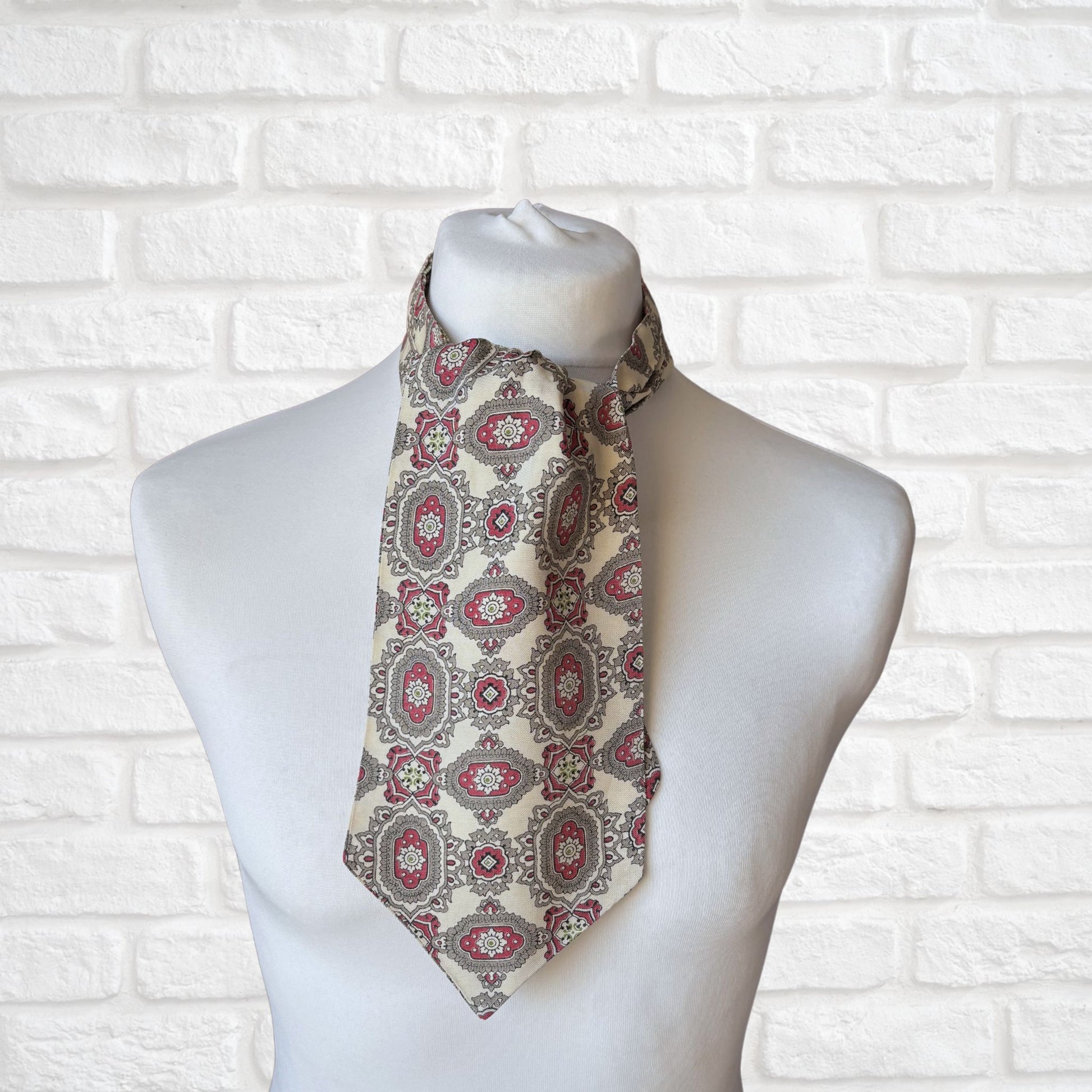 A vintage 60s mod style cream cravat with a paisley print featuring baroque style red, grey, and green patterns, displayed on a mannequin against a white brick wall background.