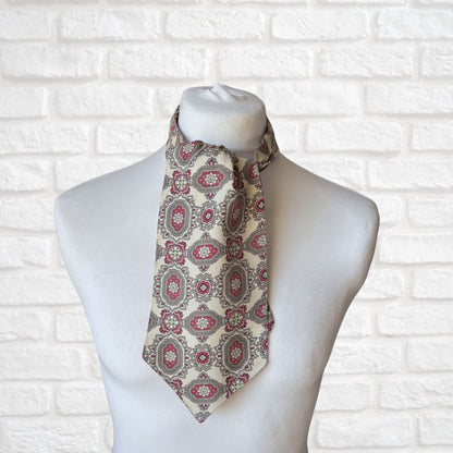 A vintage 60s mod style cream cravat with a paisley print featuring baroque style red, grey, and green patterns, displayed on a mannequin against a white brick wall background.