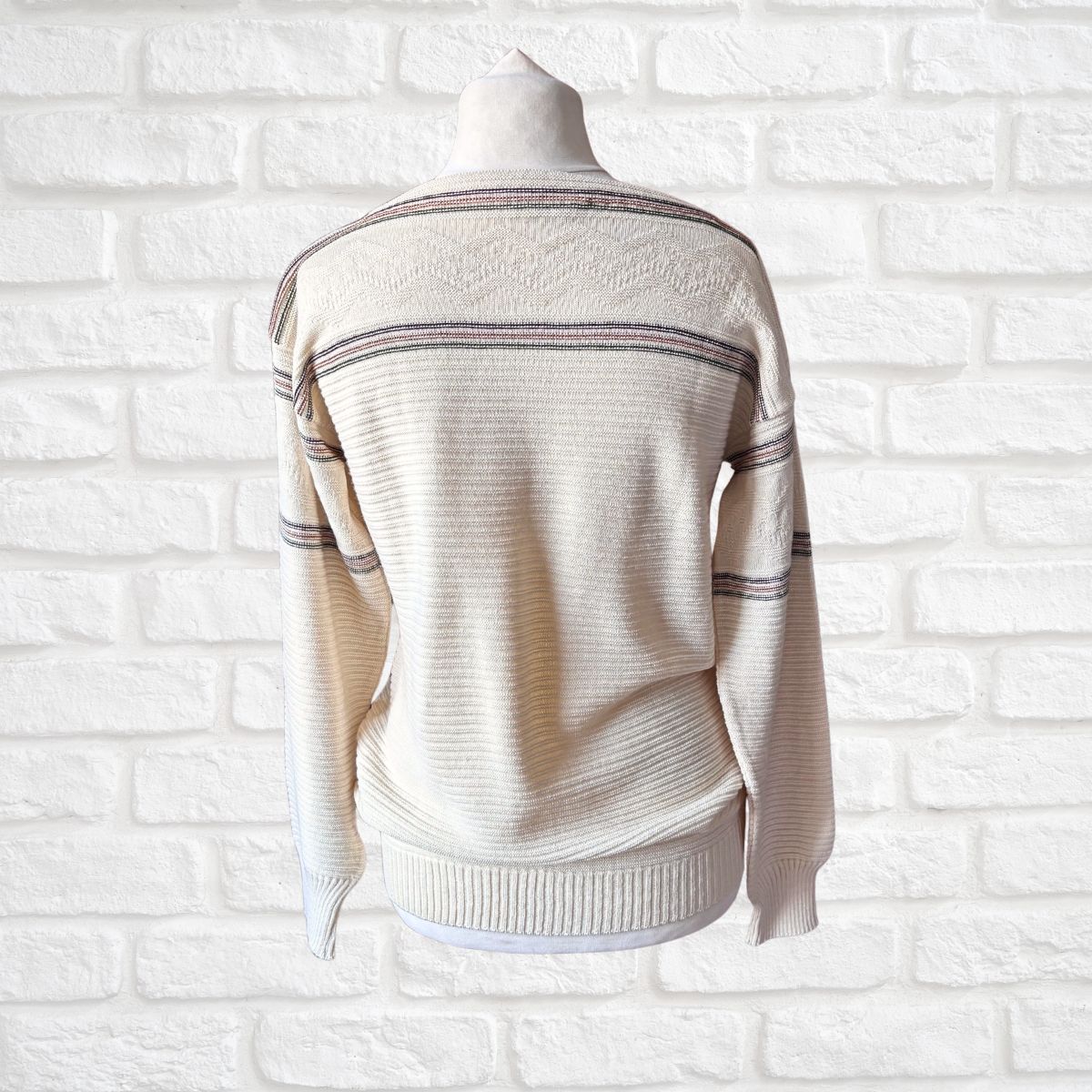A cream-colored knit sweater with thin horizontal stripes displayed on a mannequin torso against a white brick wall background