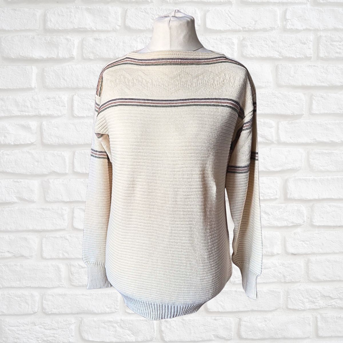 A cream-colored ribbed knit sweater with thin horizontal stripes, displayed on a mannequin torso against a white brick wall background.