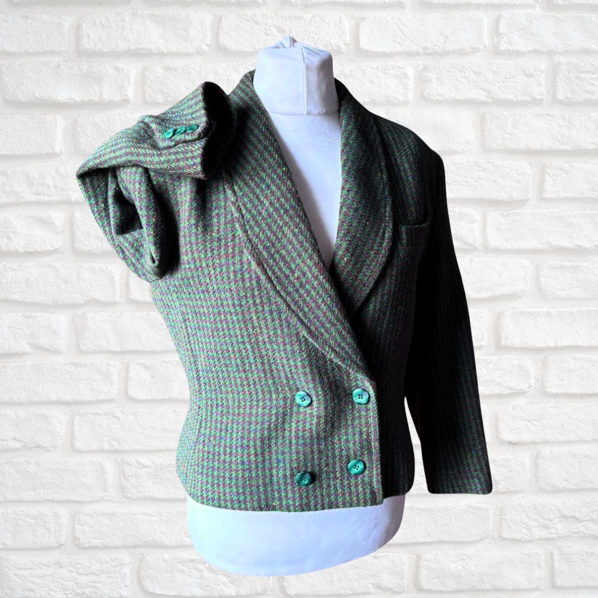 A green and brown herringbone tweed wrap jacket with turquoise buttons, displayed on a white mannequin torso against a white brick wall background.