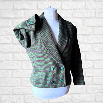 A green and brown herringbone tweed wrap jacket with turquoise buttons, displayed on a white mannequin torso against a white brick wall background.
