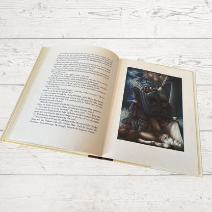 An open hardcover book laid flat on a white wood surface, showing printed text on the left page and a fantasy-style illustration on the right page.