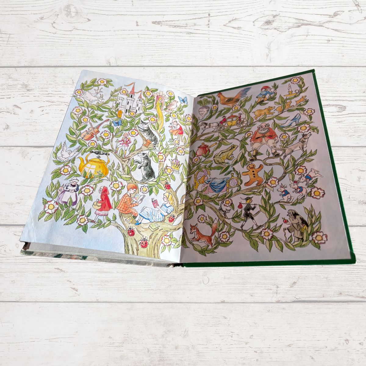 An open illustrated book or album spread on a whitewashed wooden surface, showing two illustrated tree pages filled with colorful animals, flowers, and fantasy creatures.