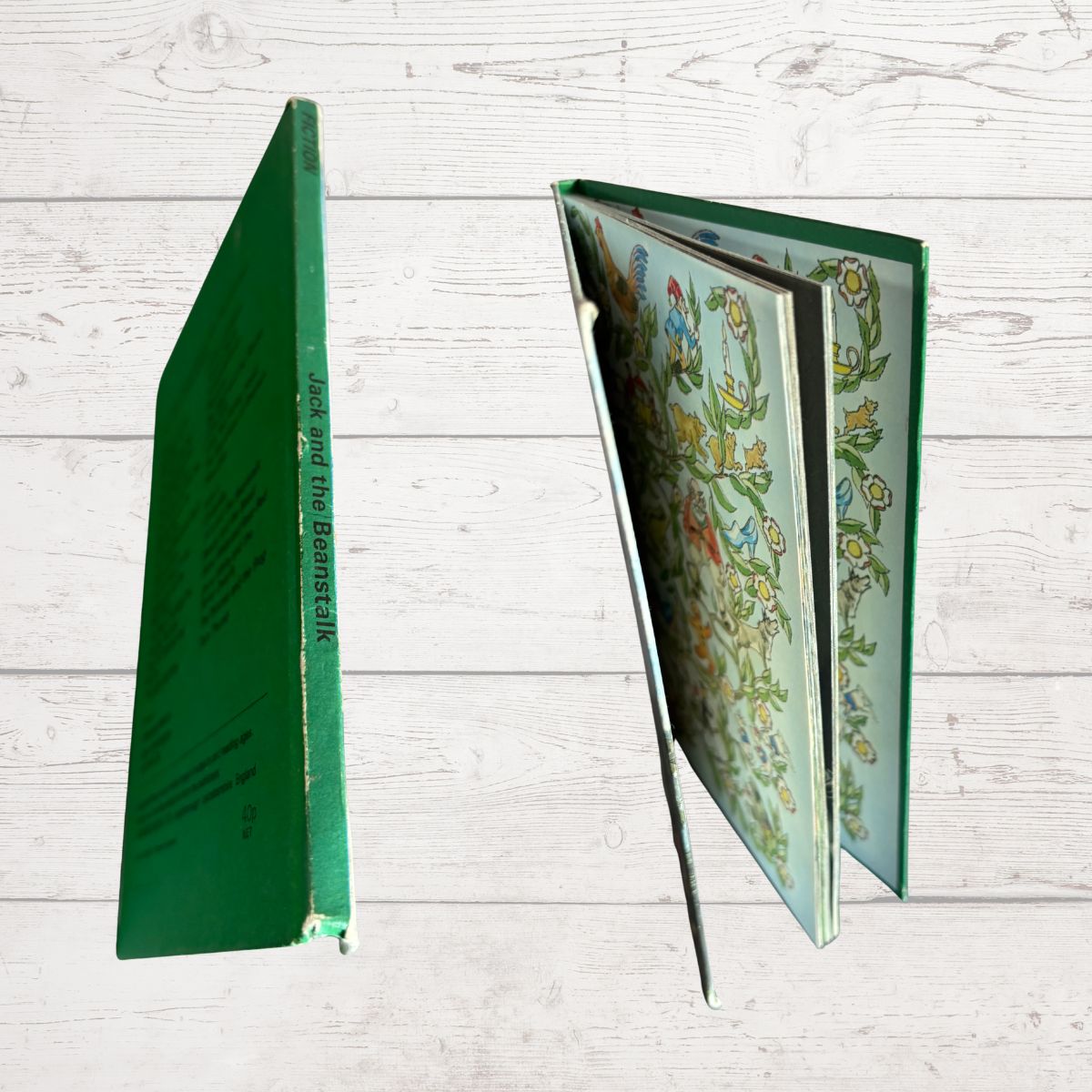 A hardcover book shown from multiple angles on a white wood plank background. One view shows the green spine with vertical title text, and the other shows the open floral-patterned interior pages.