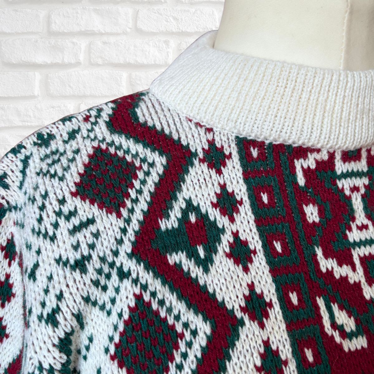 A close-up of a knit sweater on a mannequin torso, featuring a white ribbed mock neck and a Fair Isle pattern in dark green and deep red on a white base, set against a white brick wall background.
