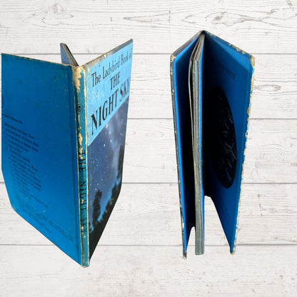 Vintage hardcover book titled "The Labeled Book of The Night Sky" shown open to display the blue cover, spine, and interior pages on a white wood plank background.