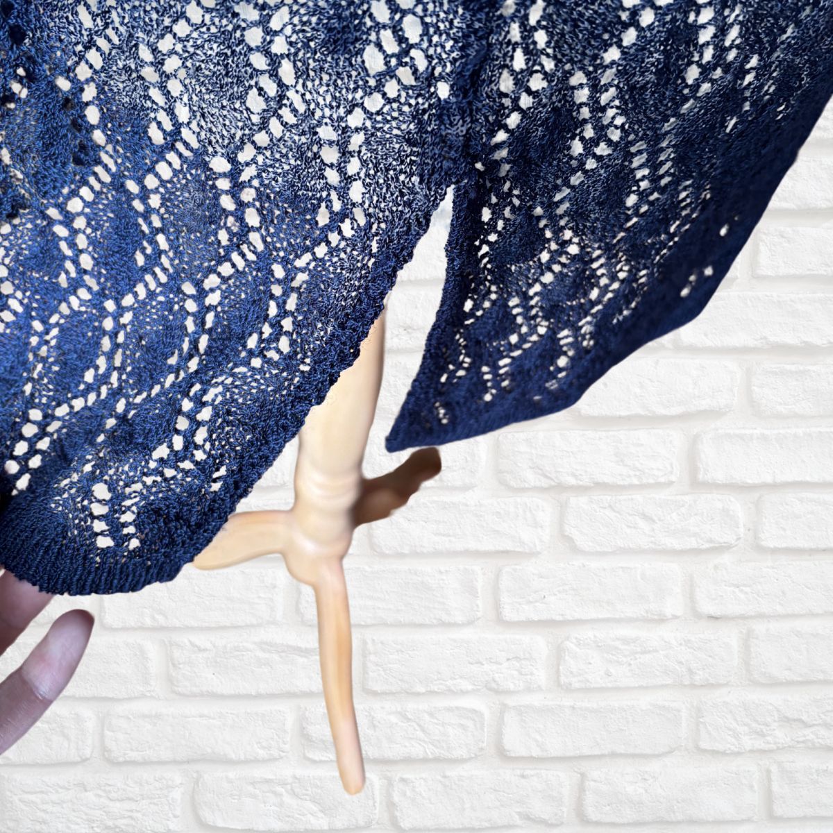 A navy blue open-knit lace waistcoat draped over a light wood hanger against a white brick wall background.