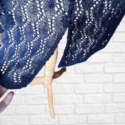 A navy blue open-knit lace waistcoat draped over a light wood hanger against a white brick wall background.