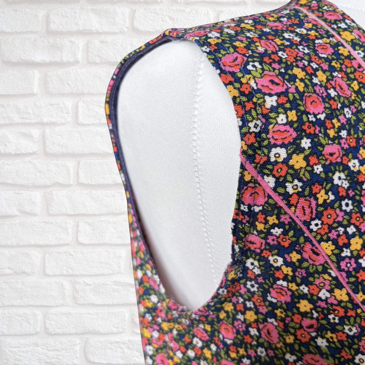 Close-up of a sleeveless floral dress on a white mannequin torso, showing a colourful print with pink, yellow, orange, and white flowers on a dark background, against a white brick wall.