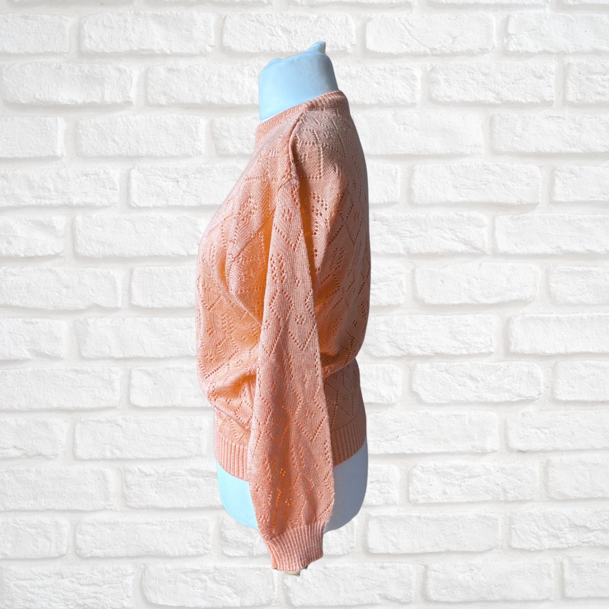 A peach-colored open-knit sweater displayed on a light blue mannequin torso, set against a white painted brick wall background.