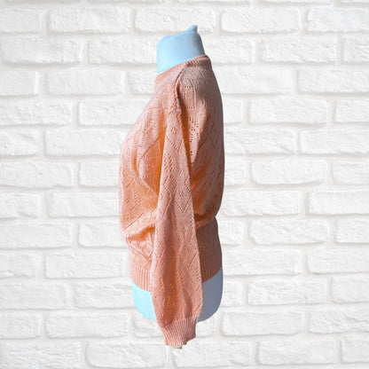 A peach-colored open-knit sweater displayed on a light blue mannequin torso, set against a white painted brick wall background.