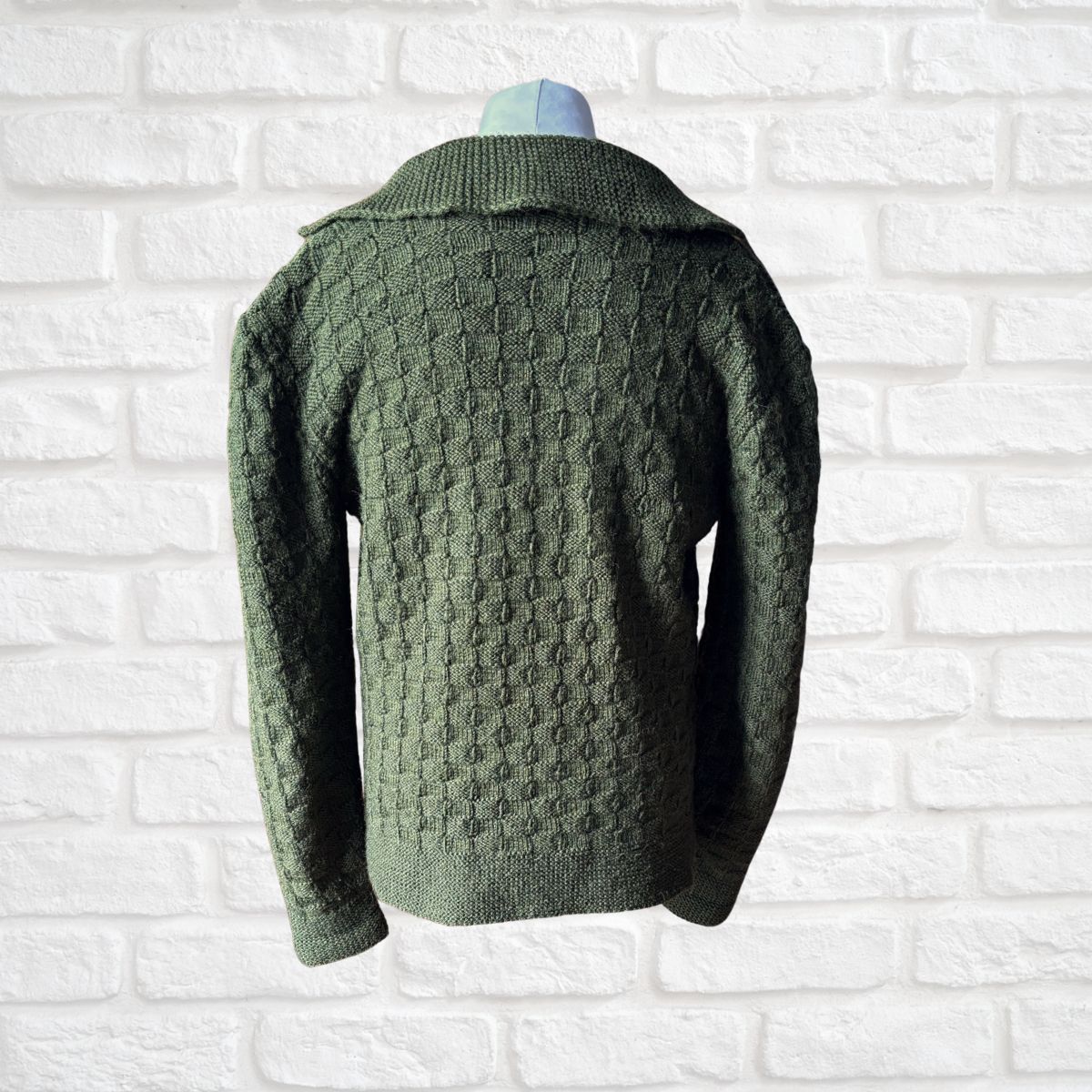 A dark green textured knit sweater displayed on a mannequin torso against a white brick wall background.