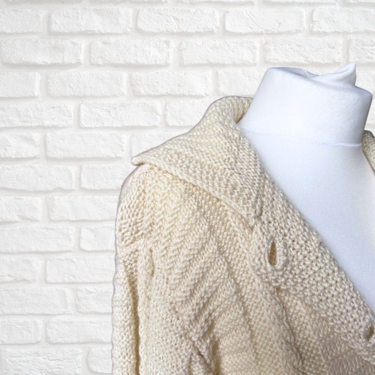 A cream-colored chunky knit cardigan sweater draped on a white mannequin torso, set against a white painted brick wall background.