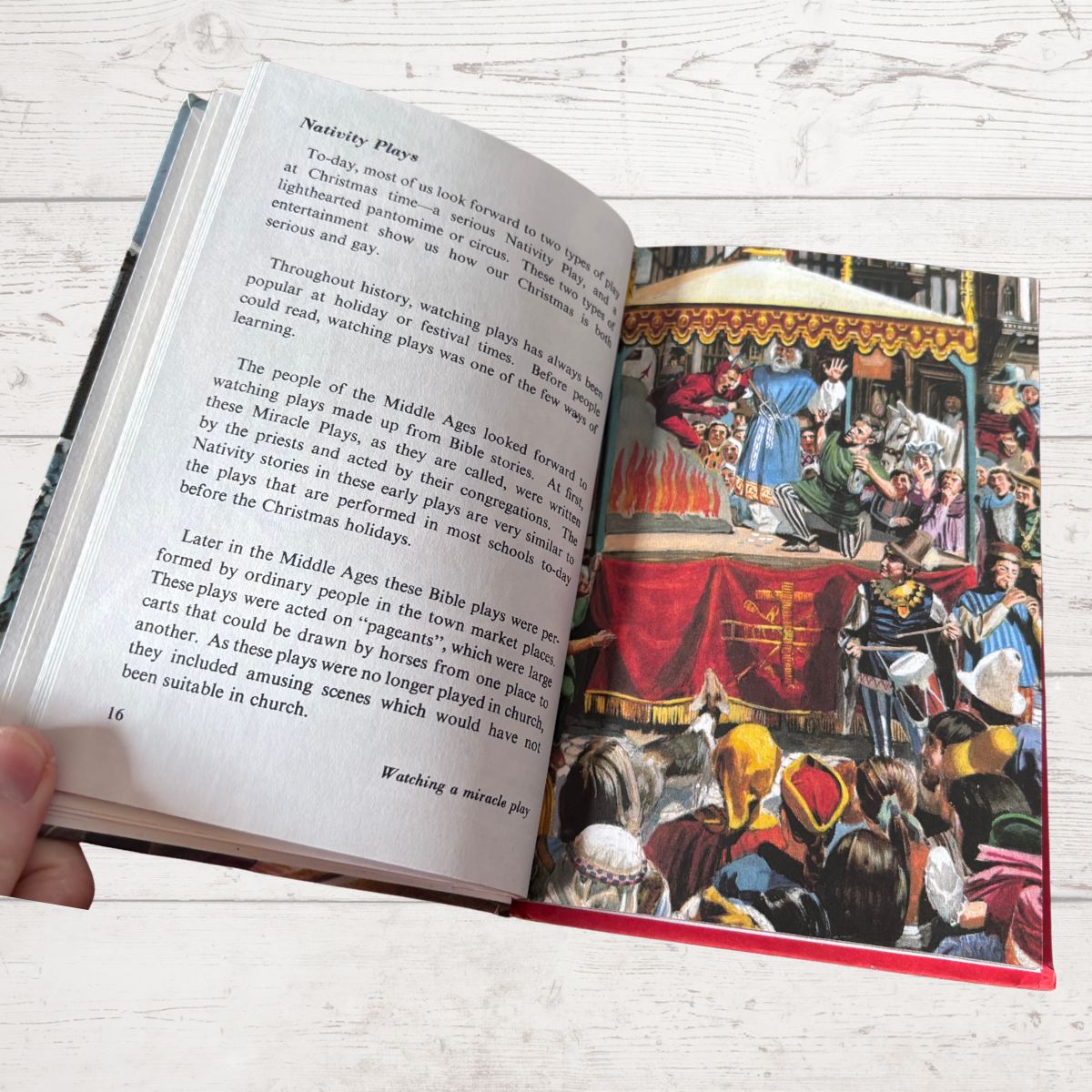 An open book being held by a hand, showing printed text on one page and a colorful illustration of a festive medieval scene on the other. The book is photographed on a light whitewashed wood surface.