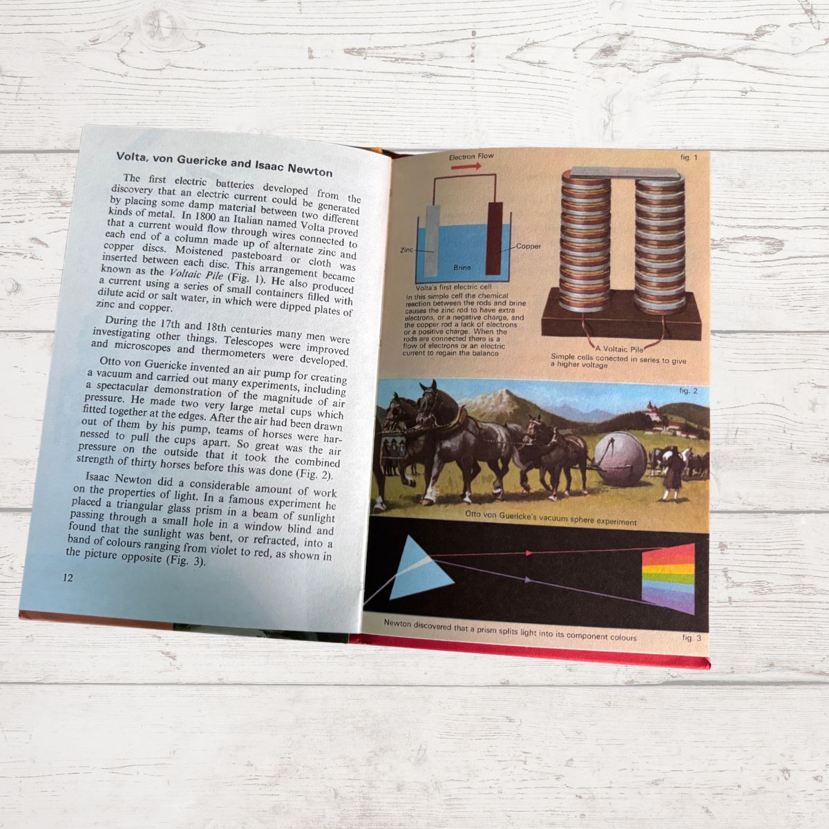 An open vintage book showing pages about Volta, van Quirkle, and Isaac Newton, with diagrams and small historical photographs, photographed on a light wood surface.