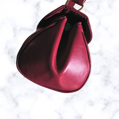 A close-up top-down view of a deep red leather pouch or small bag with a gathered, drawstring-like top edge, set against a white marble background.