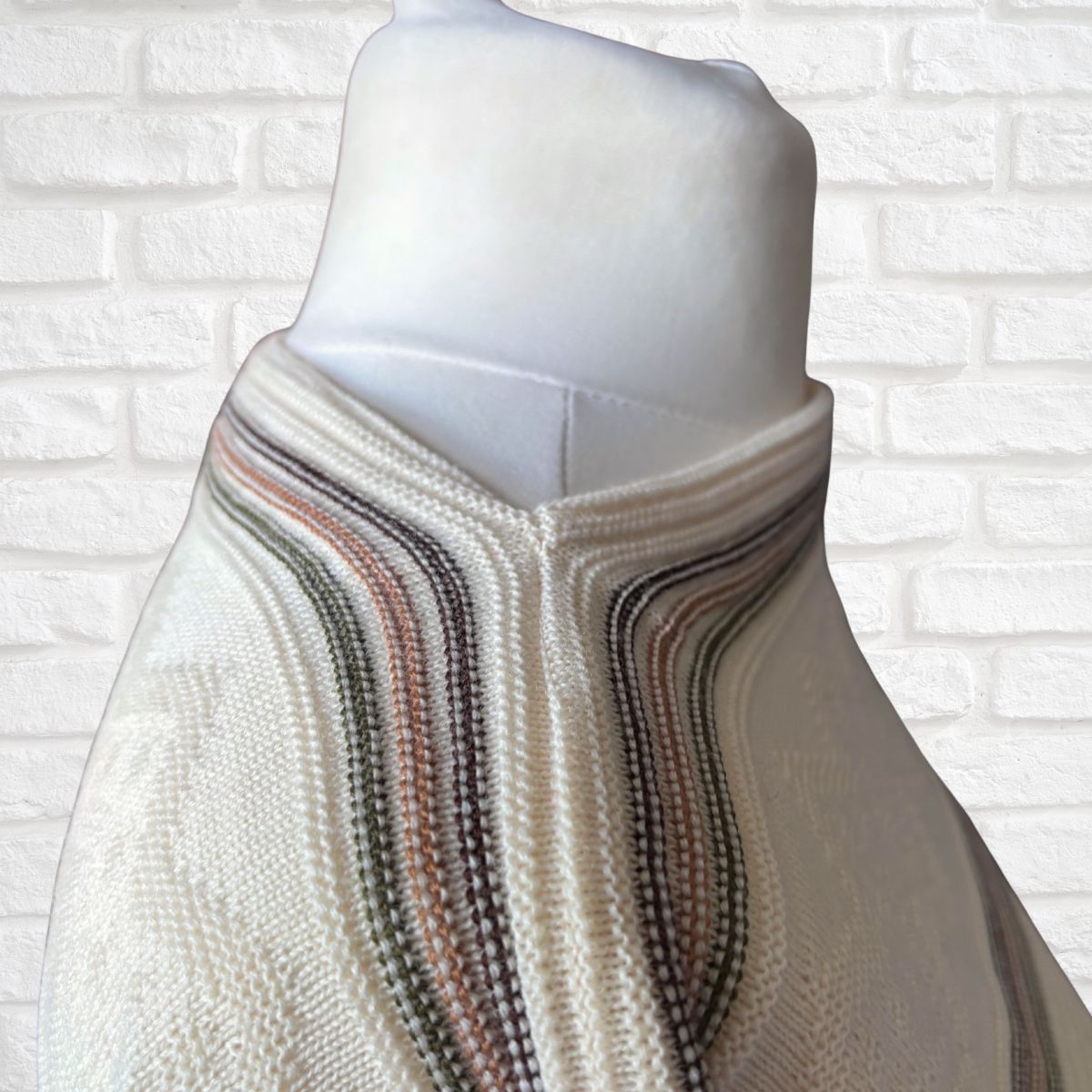 A cream-colored knit sweater with wavy stripes in muted brown, green displayed on a white mannequin torso against a white brick wall background.