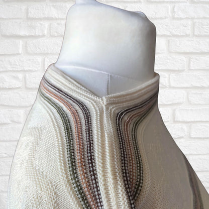 A cream-colored knit sweater with wavy stripes in muted brown, green displayed on a white mannequin torso against a white brick wall background.