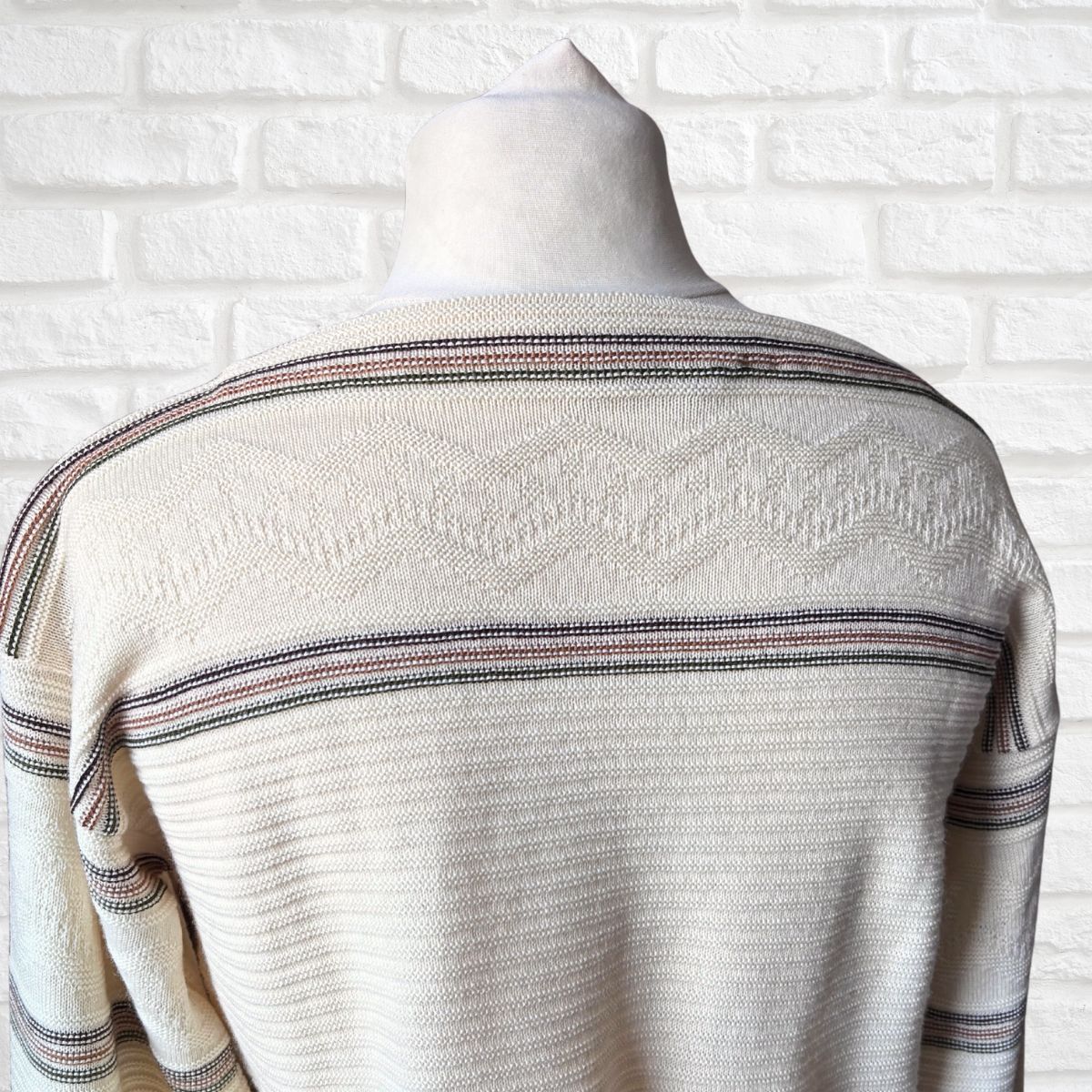 A cream-colored knit sweater with horizontal stripes  on a mannequin torso against a white brick wall background.