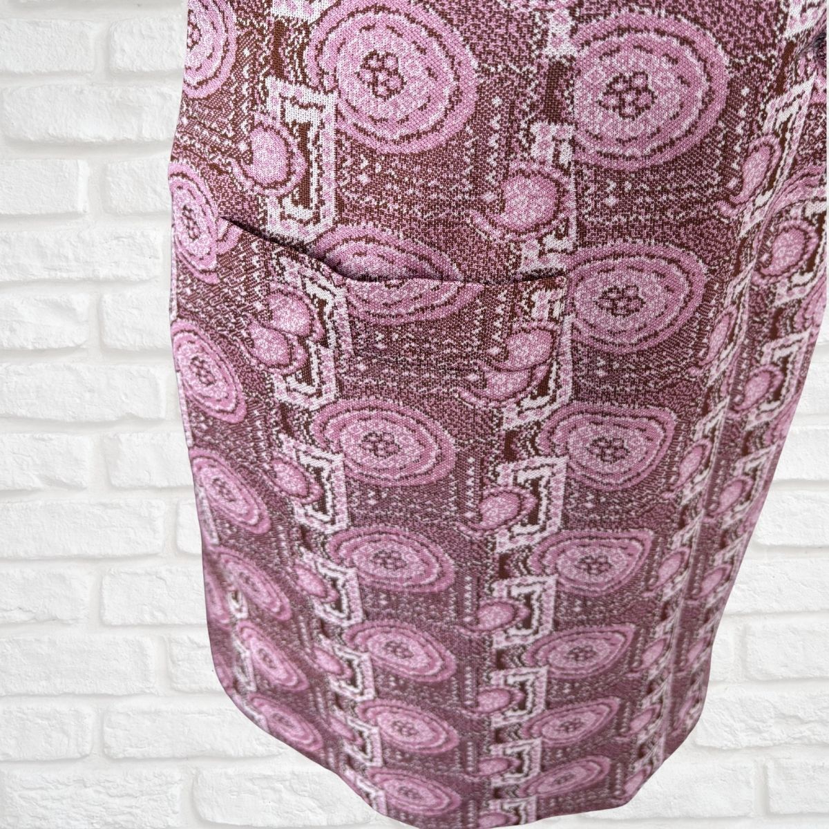 A close-up of a patterned fabric dress featuring a dusty rose and pink paisley design against a white brick wall background.