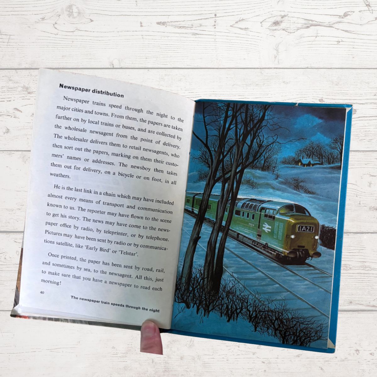 An open vintage children's book showing a two-page spread with printed text on the left page and a colorful illustration of a green train traveling through a forest on the right page, held open by a hand against a white wood background.