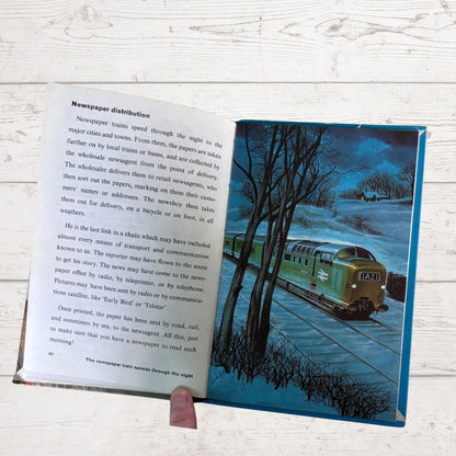 An open vintage children's book showing a two-page spread with printed text on the left page and a colorful illustration of a green train traveling through a forest on the right page, held open by a hand against a white wood background.
