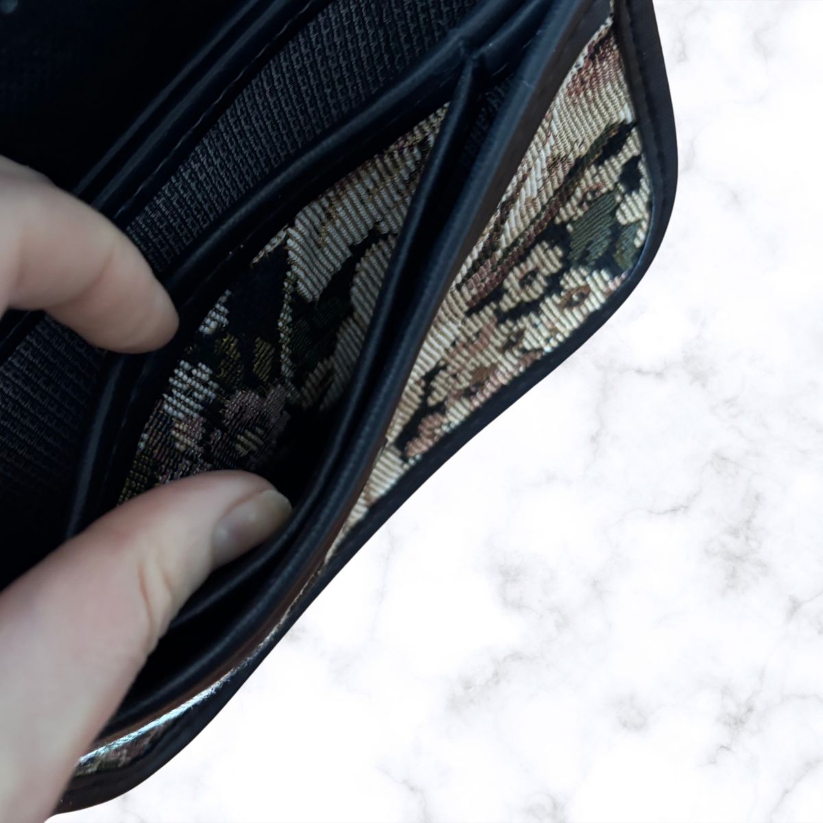 A close-up of a black bag being opened by a hand, showing patterned interior card slots on a white marble surface.