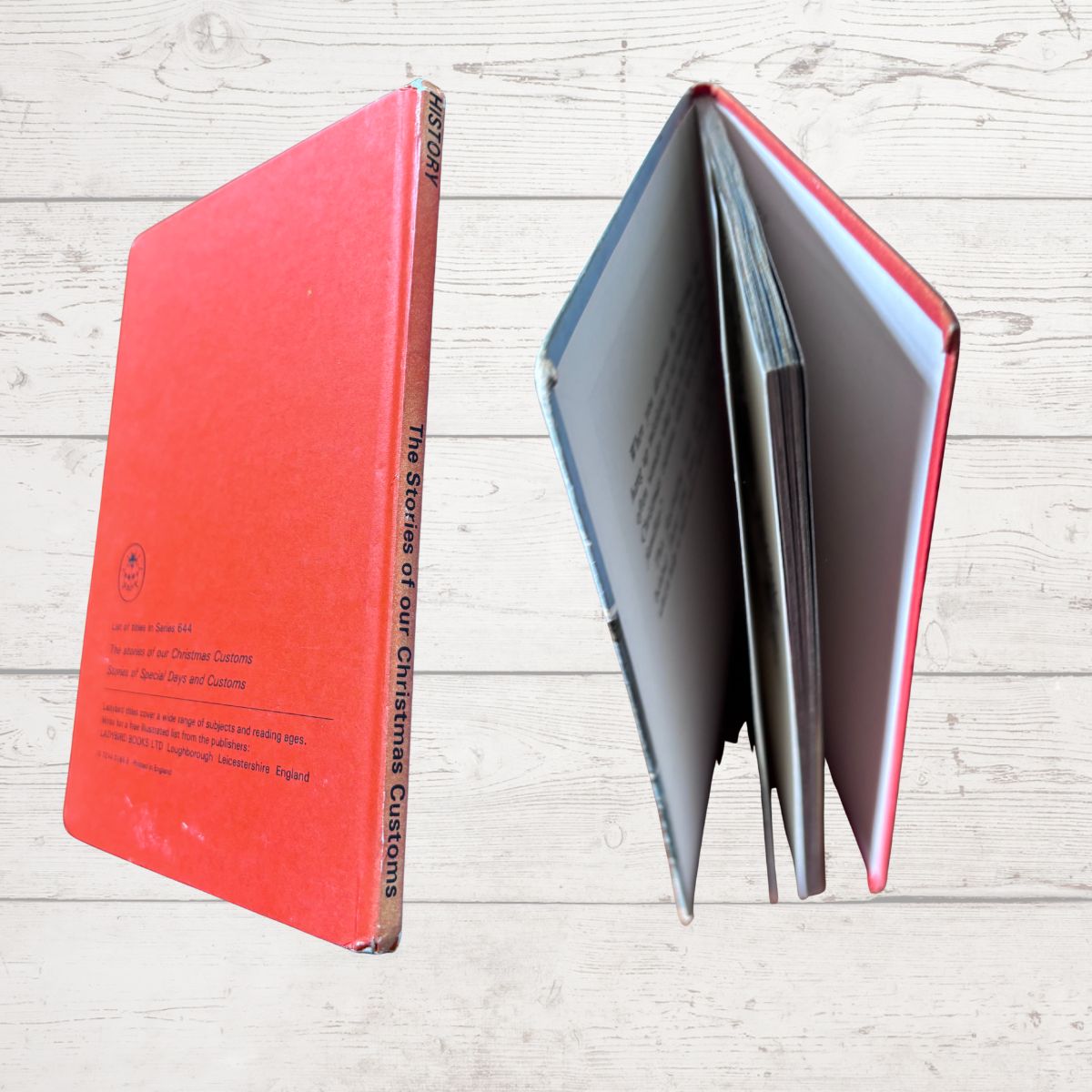 A vintage hardcover book titled 'The Stories of Our Christmas Customs ' shown standing upright next to an open view of the same book, revealing the interior pages and red binding, set against a light wood plank background 