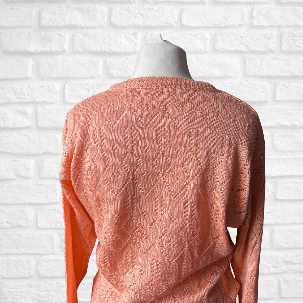 A peach-colored knit sweater with an intricate openwork diamond pattern, displayed on a mannequin torso against a white brick wall background.