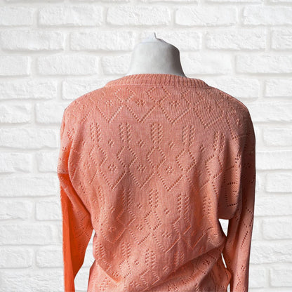 A peach-colored knit sweater with an intricate openwork diamond pattern, displayed on a mannequin torso against a white brick wall background.
