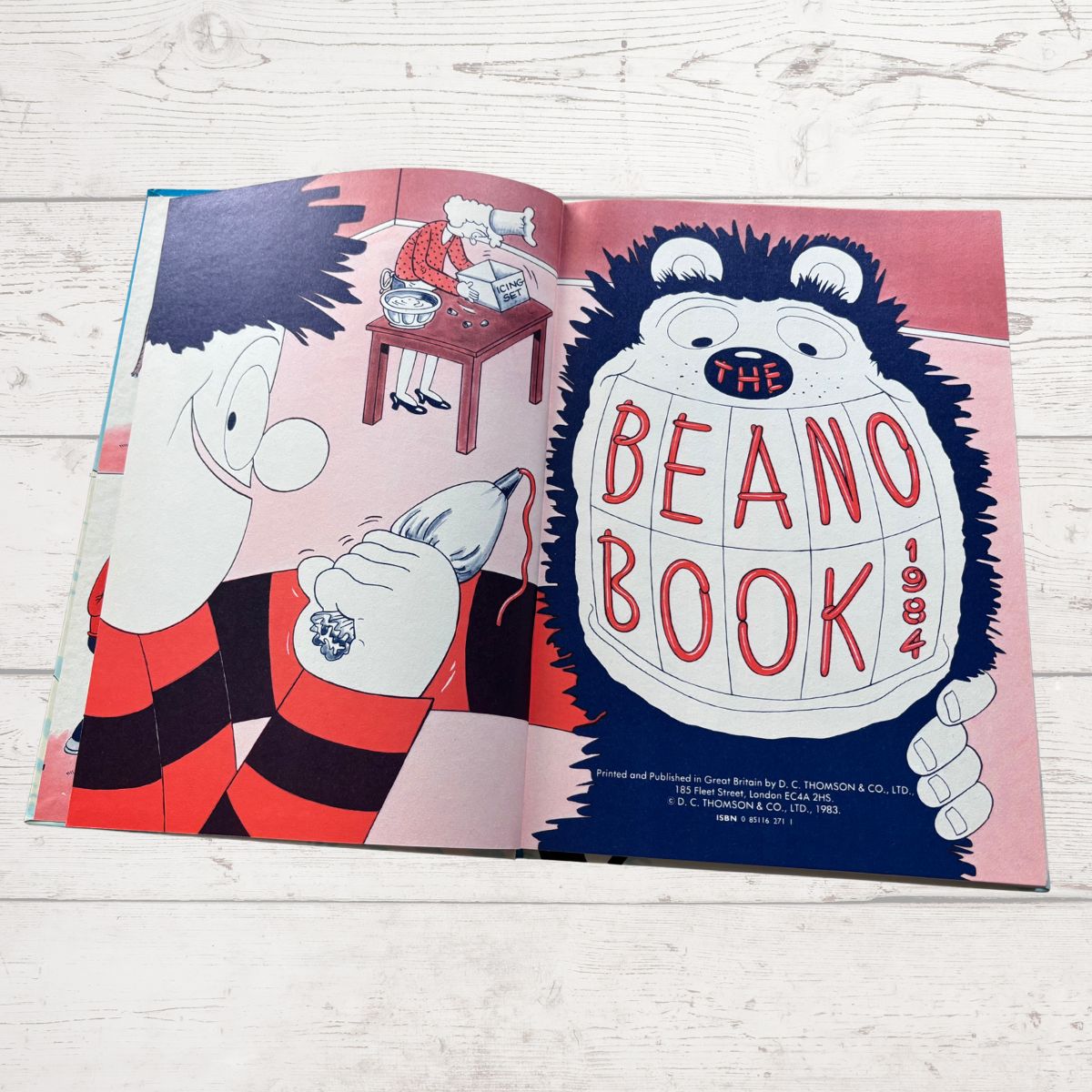 An open illustrated book titled 'The Beano Book 1964' laid flat on a light wood surface. The pages show cartoon characters and bold red lettering on a pink and blue background.