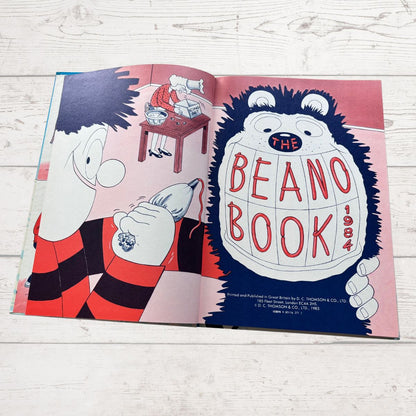 An open illustrated book titled 'The Beano Book 1964' laid flat on a light wood surface. The pages show cartoon characters and bold red lettering on a pink and blue background.