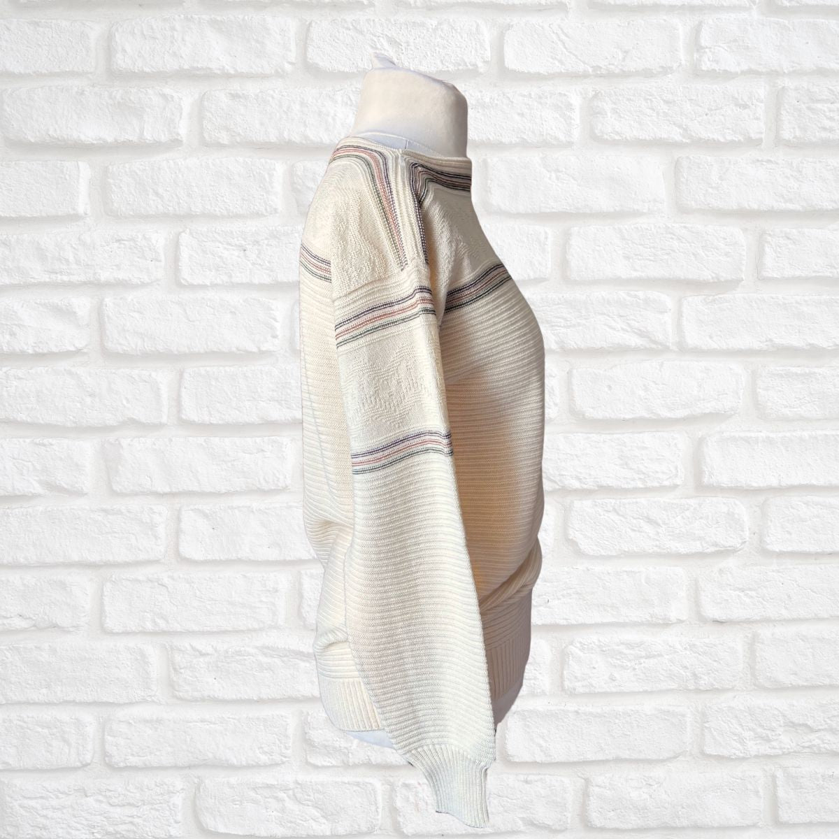 A cream-colored knit sweater with thin horizontal stripes in green and brown, displayed on a mannequin torso against a white brick wall background.