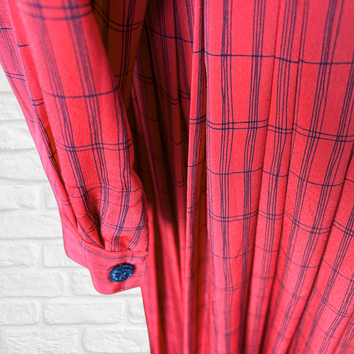 A close-up of a red plaid dress with black lines, featuring a buttoned cuff and a white brick wall background.