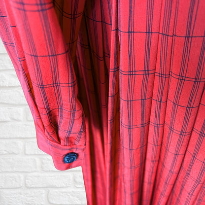 A close-up of a red plaid dress with black lines, featuring a buttoned cuff and a white brick wall background.