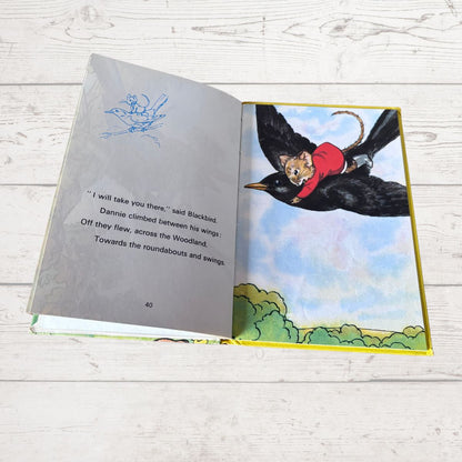 An open children's book lying on a white wood surface, showing a printed illustration of a cat riding a blackbird in flight on one page and text on the other page.