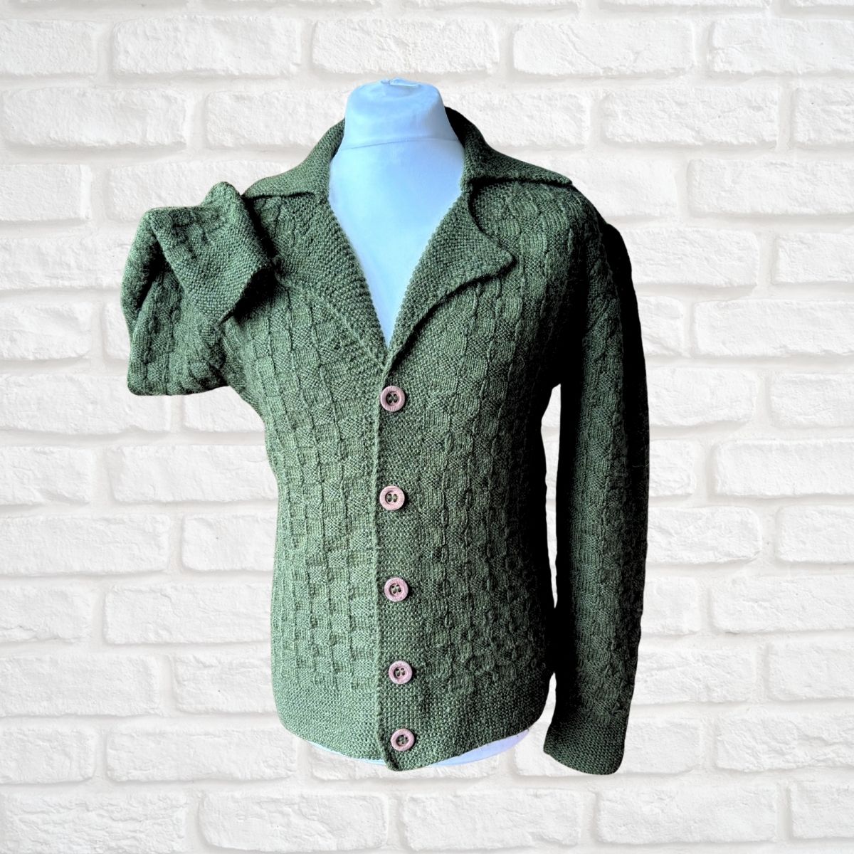 A green cable-knit cardigan sweater with a collar and pink buttons, displayed on a light blue mannequin torso against a white brick wall background.